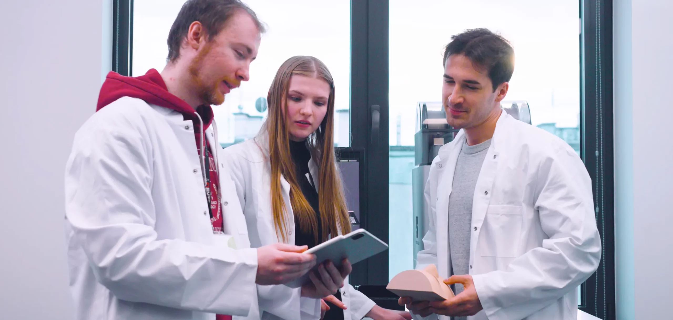 Three researchers, dressed in white lab coats, are engaged in a discussion in a bright, modern laboratory. One researcher, wearing a red hoodie under his coat, holds a tablet and explains something to the other two. A female researcher with long blonde hair listens attentively, while the third researcher, holding a model, nods in agreement. Large windows in the background let in ample natural light, highlighting the collaborative and innovative atmosphere of the lab. Three researchers, dressed in white lab coats, are engaged in a discussion in a bright, modern laboratory. One researcher, wearing a red hoodie under his coat, holds a tablet and explains something to the other two. A female researcher with long blonde hair listens attentively, while the third researcher, holding a model, nods in agreement. Large windows in the background let in ample natural light, highlighting the collaborative and innovative atmosphere of the lab.</p><p>