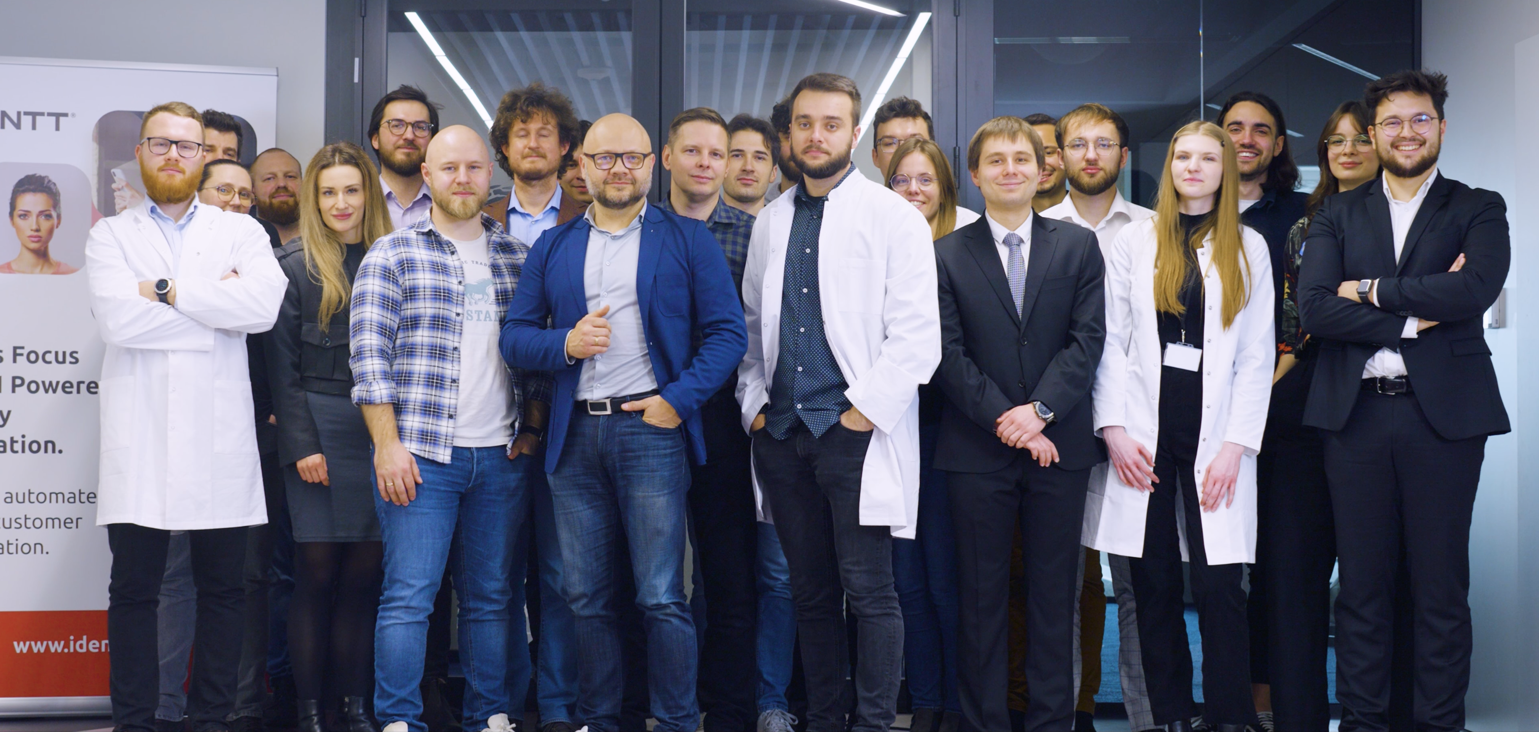 A diverse group of professionals, consisting of researchers and office staff, stands together for a group photo in a modern office setting. Some members are wearing lab coats, while others are dressed in business casual attire or formal suits. They are posed in front of a glass door, with a banner displaying the logo and tagline of their organization visible to the left. The atmosphere is one of teamwork and professionalism, reflecting a collaborative and innovative workplace.</p><p>