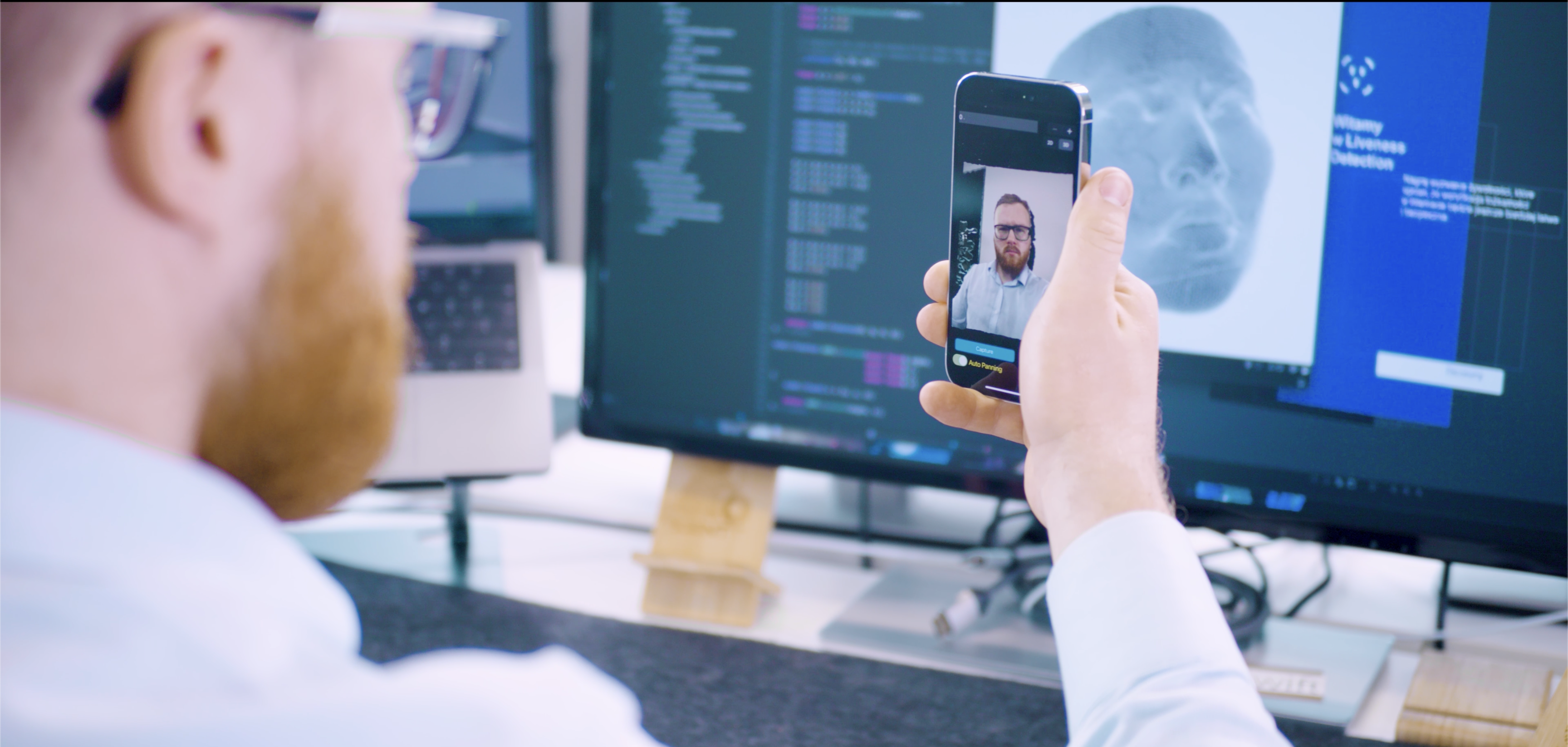 Male researcher sitting in front of a computer screen, holding iPhone with a Liveness Detection demo application showcasing depth representation of analyzed face