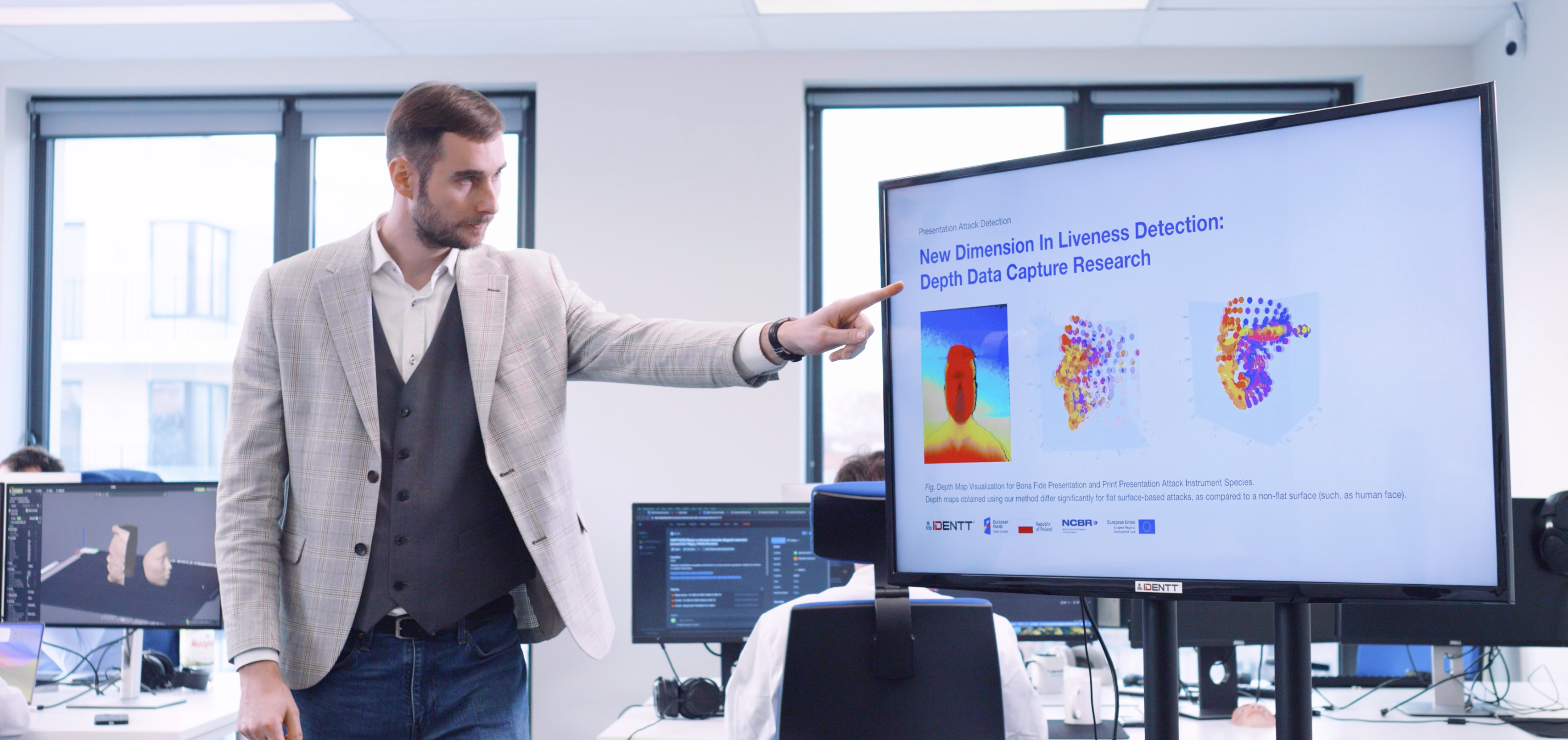 A researcher in a gray plaid blazer and white shirt is presenting his findings in a modern office. He points to a large screen displaying a presentation titled "New Dimension in Liveness Detection: Depth Data Capture Research." The screen shows various colorful data visualizations. Other employees work at their desks in the background, with computer monitors displaying related data and research. The bright office space, with large windows and a collaborative atmosphere, highlights the innovative and research-driven environment. A researcher in a gray plaid blazer and white shirt is presenting his findings in a modern office. He points to a large screen displaying a presentation titled "New Dimension in Liveness Detection: Depth Data Capture Research." The screen shows various colorful data visualizations. Other employees work at their desks in the background, with computer monitors displaying related data and research. The bright office space, with large windows and a collaborative atmosphere, highlights the innovative and research-driven environment.</p><p>