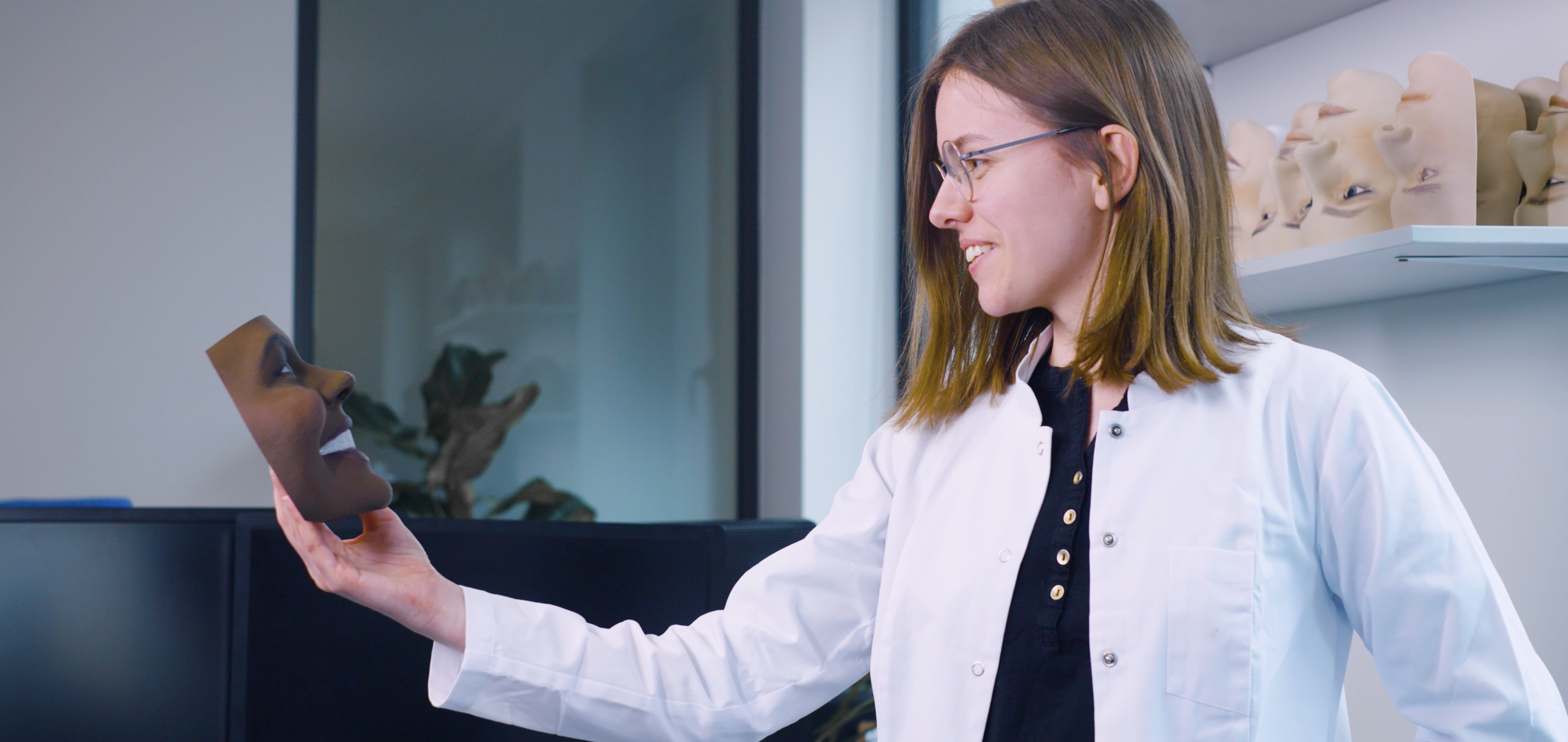 A female researcher in a white lab coat and glasses is holding and examining a detailed 3D printed model of a human face. She is smiling slightly as she looks at the model, which is brown in color. Behind her, shelves display multiple similar face models, showcasing the high-tech and innovative nature of the research being conducted in the laboratory. The setting is clean and well-organized, reflecting a professional and focused environment.</p><p>